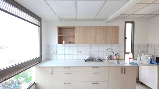 מטבח מודרני עם ארונות לבנים, אריחי קיר, מדפים עליונים מעץ וחלונות גדולים במשרד במנחם בגין; חומרי ניקוי מונחים על השיש – מושלם למשרדים להשכרה בתל אביב.