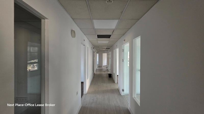 מסדרון ארוך בבניין משרדים ברחוב מנחם בגין בתל אביב, עם קירות לבנים, מספר דלתות וחלונות משני הצדדים, ואריחי תקרה – חלקם חסרים – אידיאלי להשכרת משרדים.