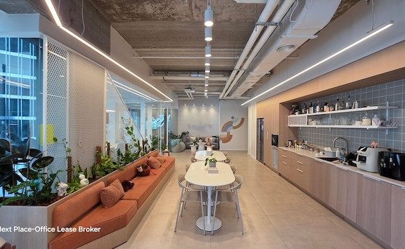 מטבח משרדי מודרני ופינת ישיבה עם שולחן ארוך, ספסלי ישיבה מובנים, צמחים, ארונות אלגנטיים ותאורה בהירה במגדל סקיי; תקרה פתוחה עם צינורות חשופים מוסיפה סגנון למשרד להשכרה זה.