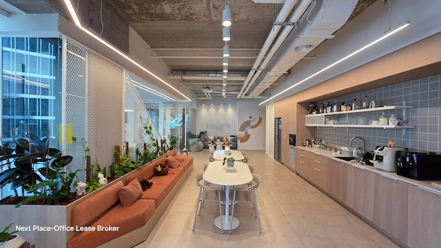 מטבח משרדי מודרני ופינת ישיבה עם שולחן ארוך, ספסלי ישיבה מובנים, צמחים, ארונות אלגנטיים ותאורה בהירה במגדל סקיי; תקרה פתוחה עם צינורות חשופים מוסיפה סגנון למשרד להשכרה זה.