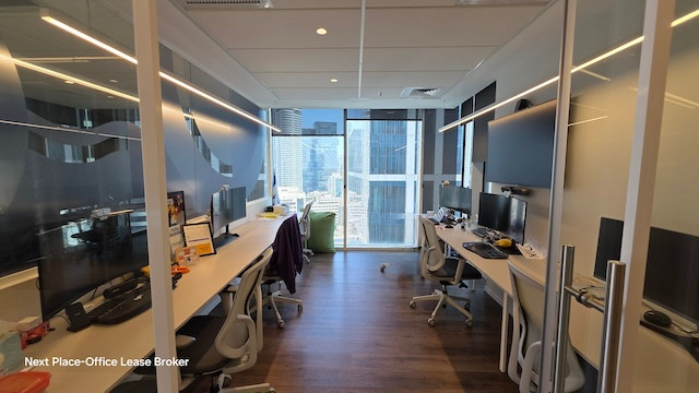 משרד מודרני להשכרה במגדל סקיי, הכולל ארבע עמדות עבודה, חלונות גדולים עם נוף לעיר, קירות זכוכית, מרפסת ומסכי מחשב על כל שולחן.