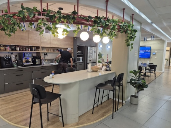 מטבח משרדי מודרני במשרד בגודל 500 מ"ר ליד בורסה רמת גן כולל אי מרכזי עם כיסאות, אדם המכין אוכל ומכשירים שונים; צמחים ירוקים תלויים מהתקרה.
