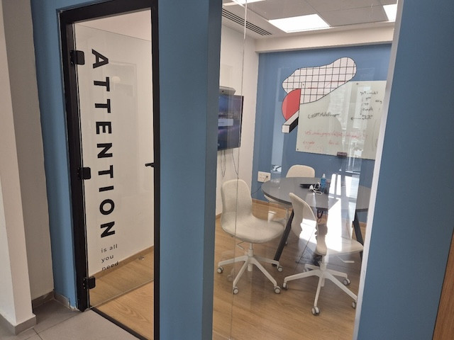 חדר ישיבות קטן ומודרני במשרד להשכרה, בורסה רמת גן, עם קירות זכוכית, ארבע כיסאות לבנים סביב שולחן עגול, מסך קיר, וטקסט מוטיבציוני על הדלת: "כל מה שאתה צריך זה תשומת לב.