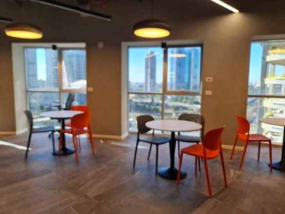 אזור ישיבה מודרני בסגנון Plug&Play עם שולחנות עגולים, כיסאות כתומים ואפורים, חלונות גדולים ונוף של בנייני העיר הנשקף מחוץ למגדל רוגובין תדהר.