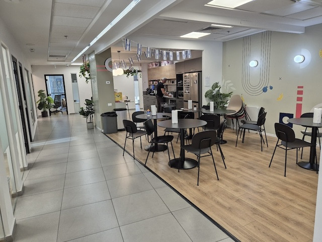 חדר מנוחה במשרד עם שולחנות עגולים וכסאות, פינת מטבח, קישוטים על הקירות ועציצים; מסדרון נראה בצד שמאל — אידיאלי עבור משרד להשכרה במשרד 500 מ"ר בבורסה רמת גן.