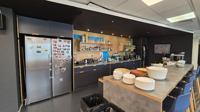 מטבח משרדי מודרני במתחם הבורסה, רמת גן, עם דלפק ארוך, קערות, מקרר נירוסטה עם תמונות, מדפים מלאים באספקה וכיסאות כחולים לאורך האי – מושלם לצרכי משרד להשכרה.