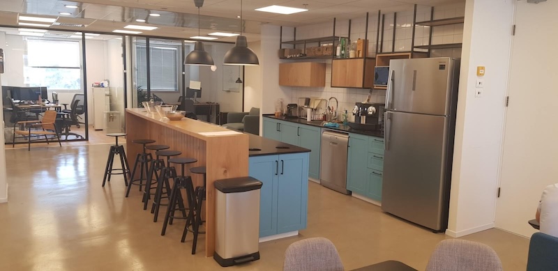 מטבח משרדי מרוהט ומודרני עם ארונות כחולים, מכשירי חשמל מנירוסטה, דלפק עץ ארוך עם שרפרפים שחורים, מנורות תלייה וחלל עבודה פתוח — משרד להשכרה אידיאלי ברחוב יגאל אלון בתל אביב.