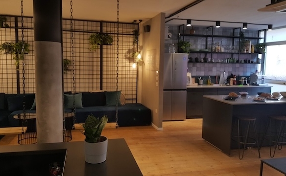 מטבח מודרני ופינת ישיבה עם כסאות בר, מקרר, צמחים ומדפים עם כלי זכוכית — עם רצפות עץ, תאורה מעורבת ואווירה מסוגננת בהשראת משרד בבית הורוד בתל אביב.