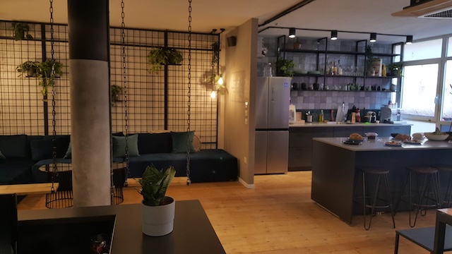 מטבח מודרני ופינת ישיבה עם כסאות בר, מקרר, צמחים ומדפים עם כלי זכוכית — עם רצפות עץ, תאורה מעורבת ואווירה מסוגננת בהשראת משרד בבית הורוד בתל אביב.