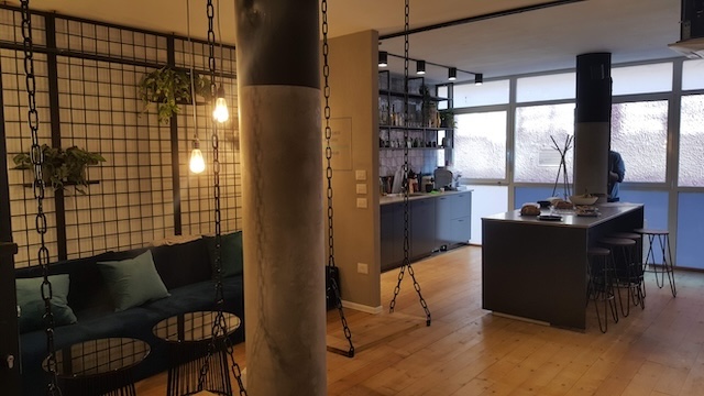 מטבח מודרני ופתוח ופינת ישיבה עם חלונות גדולים, כיסאות תלויים, מרפסת המשקיפה על תל אביב, ספה עם כריות כחולות, צמחים בתוך הבית ועיצוב בסגנון תעשייתי.