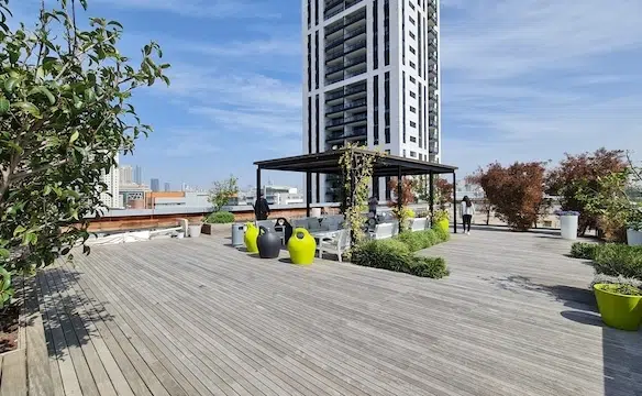 מרפסת גג מרווחת עם ריצוף עץ, עציצים ופינת ישיבה תחת פרגולה, המשקיפה על בניין מודרני גבוה ליד יגאל אלון תל אביב — מרפסת מושלמת לכל משרד להשכרה.