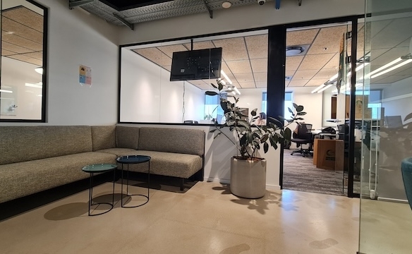 אזור המתנה מודרני למשרד עם ספה פינתית אפורה, שני שולחנות עם משטח זכוכית, עציץ ו קיר זכוכית, הממוקם במגדל משה אביב ומוצע להשכרה ברמת גן משרד מרוהט.