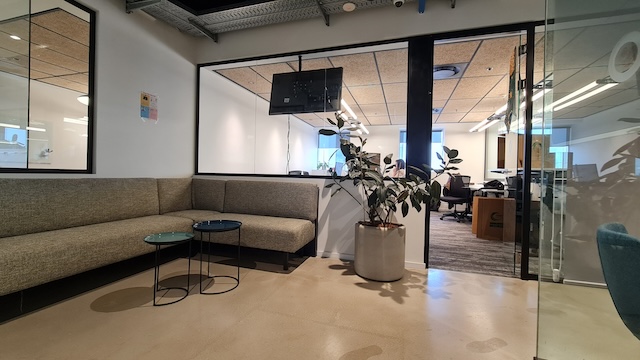 אזור המתנה מודרני למשרד עם ספה פינתית אפורה, שני שולחנות עם משטח זכוכית, עציץ ו קיר זכוכית, הממוקם במגדל משה אביב ומוצע להשכרה ברמת גן משרד מרוהט.