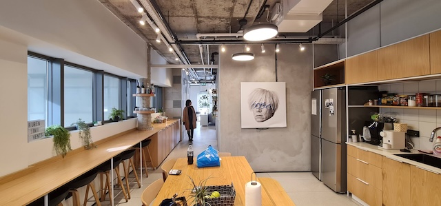 מטבח מודרני ופינת אוכל עם ריהוט מעץ, תקרה חשופה ותמונה על הקיר. מושלם למשרד במרכז תל אביב, שכן אור טבעי זורם מבעד לדלת הזכוכית בקצה החדר.