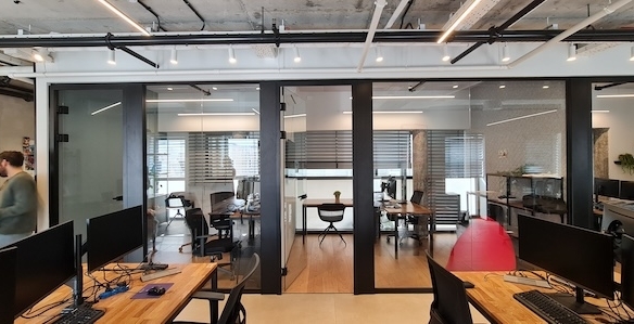 משרד להשכרה במרכז תל אביב עם חדרי ישיבות מזכוכית, שולחנות עץ עם מחשבים, ואדם הולך בצד שמאל של התמונה.