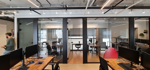 משרד להשכרה במרכז תל אביב עם חדרי ישיבות מזכוכית, שולחנות עץ עם מחשבים, ואדם הולך בצד שמאל של התמונה.