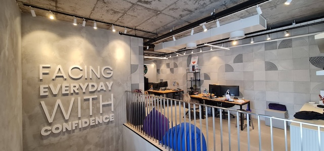 משרד מודרני עם תקרת בטון חשוף, טקסט מוטיבציוני על הקיר "להתמודד עם כל יום בביטחון", מספר שולחנות, מסכים, פופים כחולים — משרד להשכרה אידיאלי במרכז תל אביב.