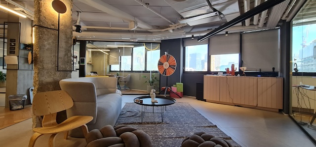 טרקלין מרוהט בסגנון מודרני עם ריהוט עכשווי, חלונות גדולים, שולחן קפה עגול וגלגל טוויה דקורטיבי ליד החלון. מבני העיר תל אביב נראים מבחוץ על דרך השלום.