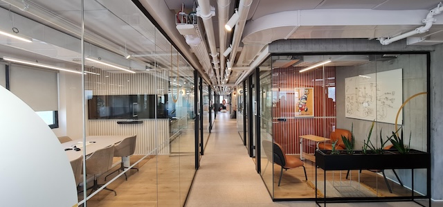 מסדרון מודרני של משרד מרוהט ברחוב דרך השלום, תל אביב, עם חדרי ישיבות בעלי קירות זכוכית משני הצדדים, כיסאות, שולחנות, צמחים, צינורות תקרה חשופים ואור טבעי בשפע.