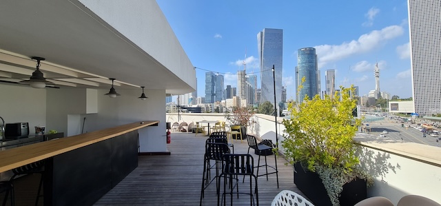 מרפסת גג בשטח של 600 מ"ר עם מקומות ישיבה בבר, שולחנות וצמחים המשקיפה על קו הרקיע של העיר עם בניינים מודרניים גבוהים תחת שמיים כחולים. אידיאלי למשרד מרוהט בדרך השלום בתל אביב.