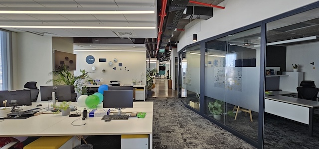 שטח משרדי מודרני בגודל 600 מ"ר להשכרה במגדל אטריום, הכולל שולחנות, מחשבים, בלונים צבעוניים, חדרי ישיבות עם קירות זכוכית, צמחים עבותים ותאורה טבעית שופעת הנכנסת מבעד לחלונות גדולים בצד שמאל.