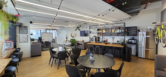 מטבח ומרחב מנוחה מודרניים במגדל אטריום, הכוללים שולחנות, כיסאות, כסאות בר, צמחים, מדפים עם פריטי מטבח, מקרר ואופניים התלויים על הקיר. משרד להשכרה – 600 מ"ר של עיצוב מודרני.