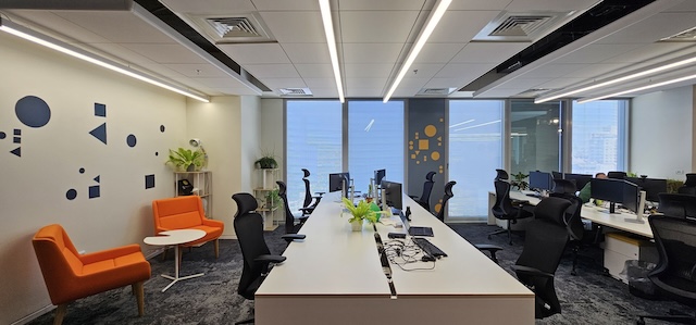 שטח משרדים מודרני להשכרה במגדל אטריום, הכולל 600 מ"ר עם שולחנות, עמדות מחשב, כיסאות שחורים, כורסאות כתומות, שולחנות עגולים, צמחים, קישוטים גיאומטריים על הקירות וחלונות גדולים ברקע.