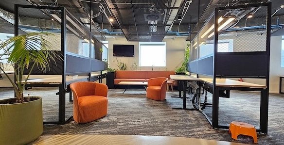 משרד מודרני עם עמדות עבודה מופרדות, כיסאות כתומים, ספה חומה, חלונות גדולים, צמחים פנימיים ותקרה עם צינורות וחשמל גלויים במגדל משה אביב ברמת גן. אידיאלי למי שמחפש משרד להשכרה יוקרתי.