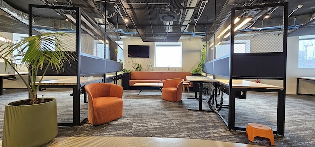 משרד מודרני עם עמדות עבודה מופרדות, כיסאות כתומים, ספה חומה, חלונות גדולים, צמחים פנימיים ותקרה עם צינורות וחשמל גלויים במגדל משה אביב ברמת גן. אידיאלי למי שמחפש משרד להשכרה יוקרתי.