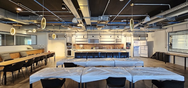 מטבח ומטבח מודרני במגדל משה אביב, עם שולחנות מכוסים בפלסטיק, תאורה תקרתית, צינורות תקרה חשופים ומכשירים שונים לאורך הקיר האחורי - מושלם למשרדים להשכרה.