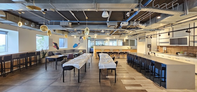 מטבח ומזנון משרדיים מודרניים ומרווחים במגדל משה אביב, עם דלפקים ארוכים, כסאות בר, שולחנות, כיסאות וצינורות חשופים בתקרה. חלק מהשולחנות מכוסים ביריעות פלסטיק – מושלם למשרד יפה או למשרד להשכרה.