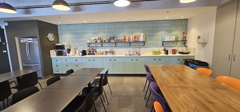 מטבח משרדי מודרני במשרד יוקרתי להשכרה, הכולל שולחנות ארוכים, כיסאות צבעוניים, מדפים עמוסים במזון ומכשירים חשמליים לאורך הקיר האחורי. ממוקם במגדל השחר, מתחם הבורסה ברמת גן.