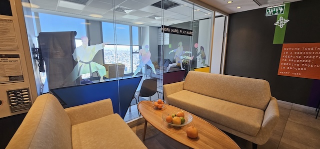 טרקלין משרדי מודרני עם ספות בצבע בז', שולחן קפה מעץ עם פירות וקיר זכוכית עם דמויות מופשטות, מציע נוף עוצר נשימה של העיר במגדל השחר, מתחם הבורסה רמת גן — מושלם למי שמחפש משרד יוקרתי להשכרה.