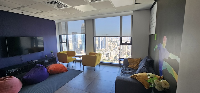 טרקלין משרדי מודרני עם פופים, כורסאות, ספה וטלוויזיה המותקנת על הקיר; חלונות גדולים המשקיפים על נוף העיר מתחם הבורסה רמת גן במגדל השחר.
