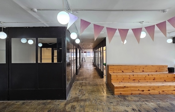 שטח משרדים מודרני להשכרה בתל אביב בבית הורוד, הכולל יציע עץ, תאורה עגולה תלויה, דגלונים ורודים, משרדים עם מסגרות זכוכית שחורות ורצפות עץ מסוגננות.