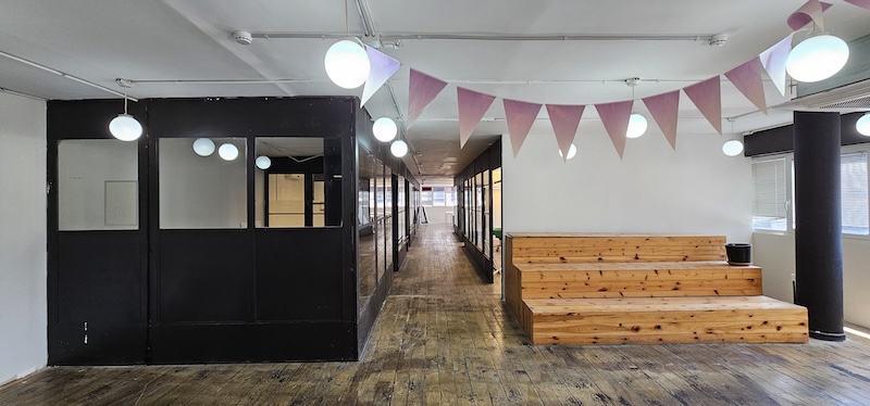 שטח משרדים מודרני להשכרה בתל אביב בבית הורוד, הכולל יציע עץ, תאורה עגולה תלויה, דגלונים ורודים, משרדים עם מסגרות זכוכית שחורות ורצפות עץ מסוגננות.