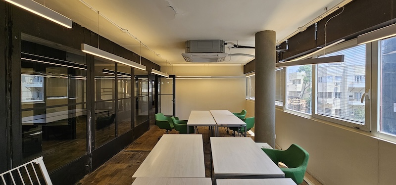 משרד להשכרה בבית הורוד בתל אביב עם חלונות גדולים, מחיצות זכוכית, שולחנות לבנים וכיסאות ירוקים, תקרה חשופה ואור טבעי.