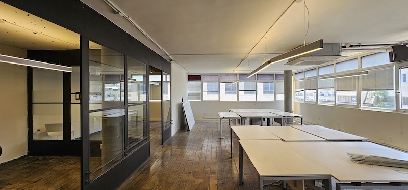 משרד מרווח להשכרה בתל אביב עם רצפות עץ, חלונות גדולים, שולחנות לבנים וחדר ישיבות עם קירות זכוכית בצד שמאל.