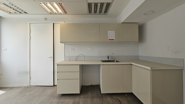 מטבח מודרני ומינימליסטי במשרד להשכרה של אגיס רבד, הכולל ארונות בצבע בז', דלת לבנה, כיור ותאורה פלורוסנטית תקרתית. החלל, ששטחו 450 מ"ר, כולל רצפת עץ וקירות אפורים בהירים למראה נקי.