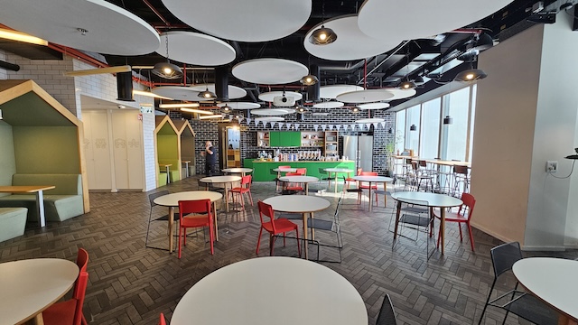 קפיטריה מודרנית בקומה 38 של מגדל אטריום עם שולחנות עגולים וכסאות אדומים, דלפק קפה ירוק בחלק האחורי, חלונות גדולים מימין ולוחות תקרה עגולים מעל הראש.