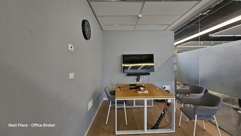 משרד להשכרה בבית קרדן, תל אביב: משרד קטן עם שולחן, ארבעה כיסאות, טלוויזיה על הקיר, שעון וכבלים גלויים; קירות זכוכית מפרידים מהאזור הסמוך.