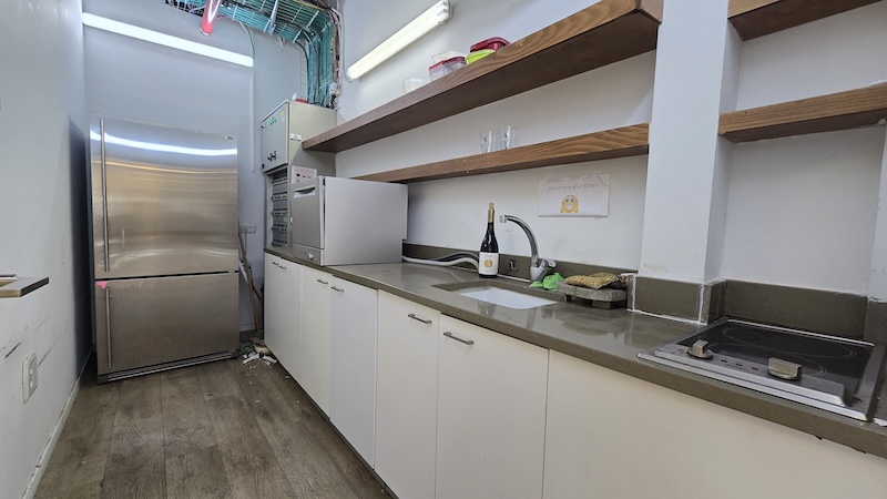 מטבחון קטן עם מקרר, מיקרוגל, מדפים פתוחים, כיור וכיריים; על השיש והמדפים מונחים פריטים שונים — מושלם למשרד שלך במתחם הבורסה.