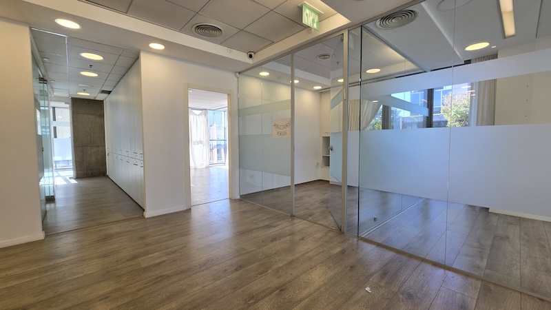 משרד מודרני עם חדרי ישיבות בעלי קירות זכוכית, ריצוף עץ, קירות לבנים ותאורת תקרה. אור השמש נכנס מבעד לחלונות ברקע, ומאפשר הצצה למרפסת הענקית במתחם הבורסה.