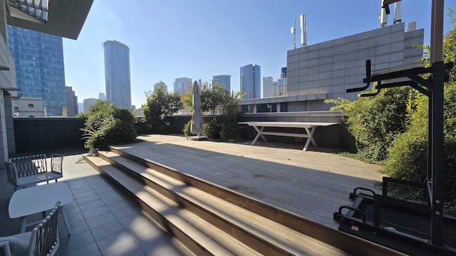 מרפסת ענקית על גג משרד במתחם הבורסה, עם דק עץ, ריהוט חוץ, ציוד כושר, צמחייה ונוף לעיר ביום שמשי.