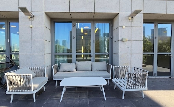 פטיו חיצוני בבית גרניט, הכולל ספה לבנה, שני כורסאות תואמות ושולחן קפה המוצב על רצפת אבן מול דלתות זכוכית — מושלם למנוחה מחוץ למשרד במתחם הבורסה.