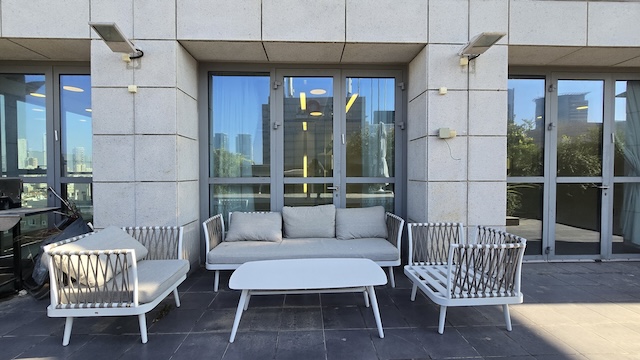 פטיו חיצוני בבית גרניט, הכולל ספה לבנה, שני כורסאות תואמות ושולחן קפה המוצב על רצפת אבן מול דלתות זכוכית — מושלם למנוחה מחוץ למשרד במתחם הבורסה.