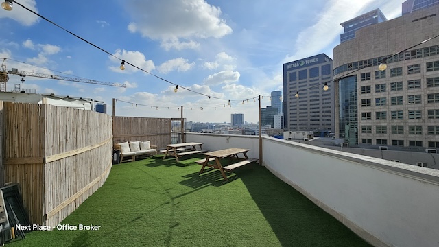 מרפסת גג עם דשא מלאכותי, ספסלי עץ, אורות מחרוזת ובנייני העיר ברקע תחת שמיים כחולים, הממוקמת ליד מנחם בגין בתל אביב — מושלמת למשרד להשכרה.