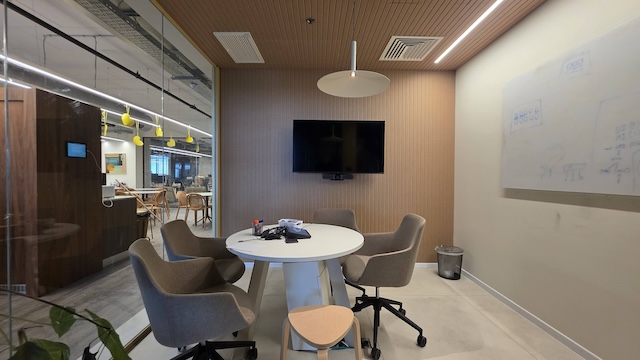 חדר ישיבות קטן עם שולחן עגול, ארבעה כיסאות, טלוויזיה, לוח לבן, טלפון וקיר זכוכית המשקיף על אזור משרדים פתוח במשרד יוקרתי להשכרה במגדלי דה וינצ'י, תל אביב.