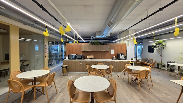 חדר מנוחה מודרני במשרד עם שולחנות עגולים, כיסאות עץ, פינת מטבח, מכונות קפה וקישוטים צהובים התלויים מהתקרה במשרד להשכרה במגדלי דה וינצ'י, תל אביב.