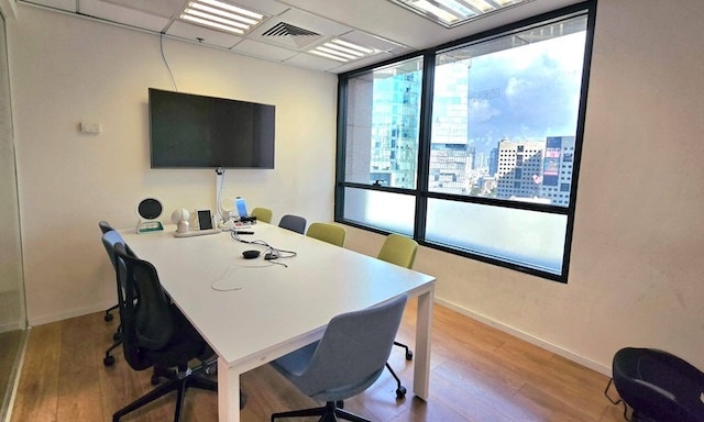 חדר ישיבות מודרני בבית שאפ במתחם הבורסה רמת גן עם שולחן לבן, שישה כיסאות, מסך קיר וחלונות גדולים המשקיפים על בנייני העיר — מושלם כמשרד להשכרה.